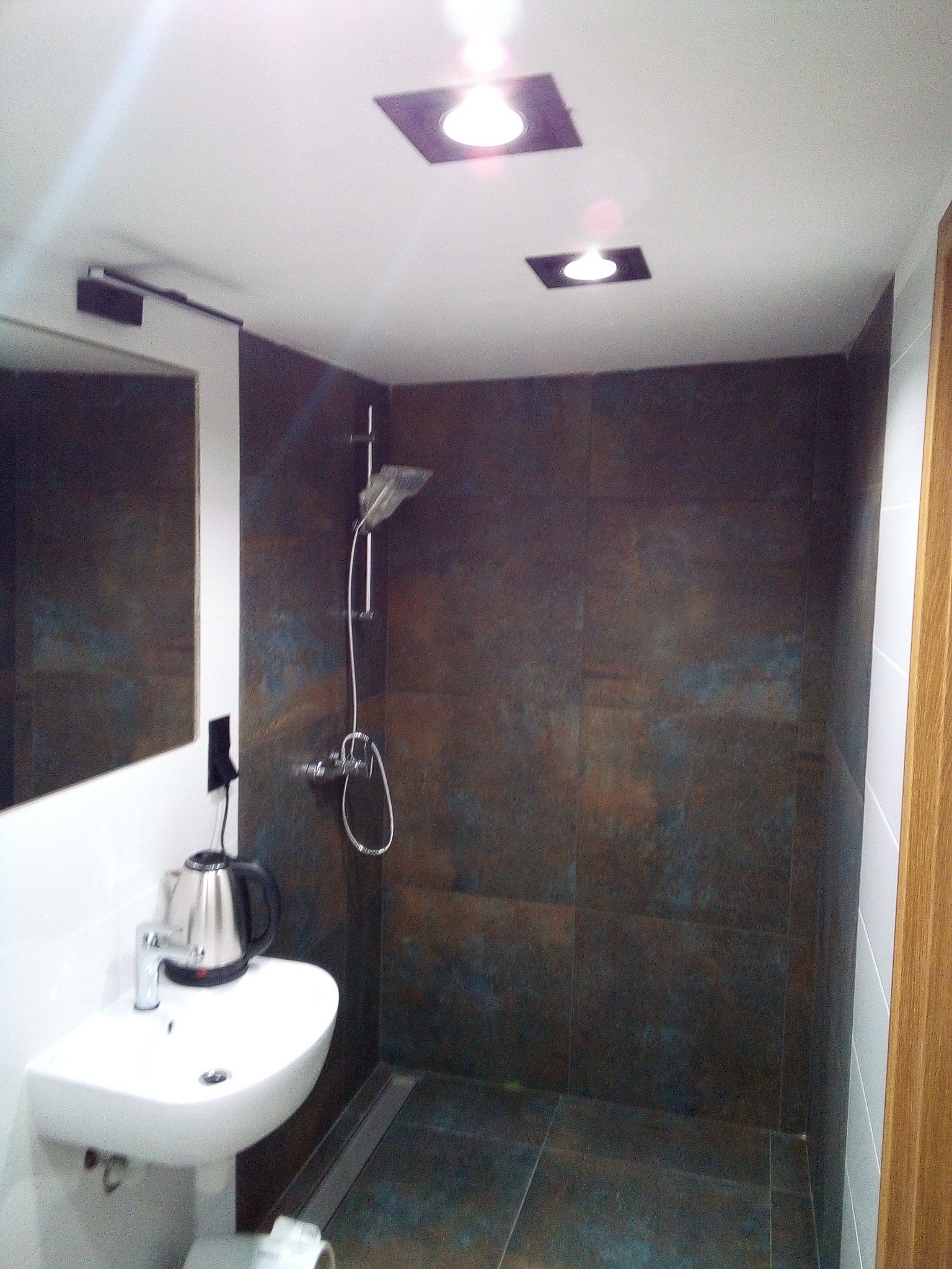 Nowoczesna łazienka z ciemnymi, metalicznymi płytkami, białą umywalką na postumencie, srebrnym czajnikiem elektrycznym i minimalistycznym oświetleniem punktowym.