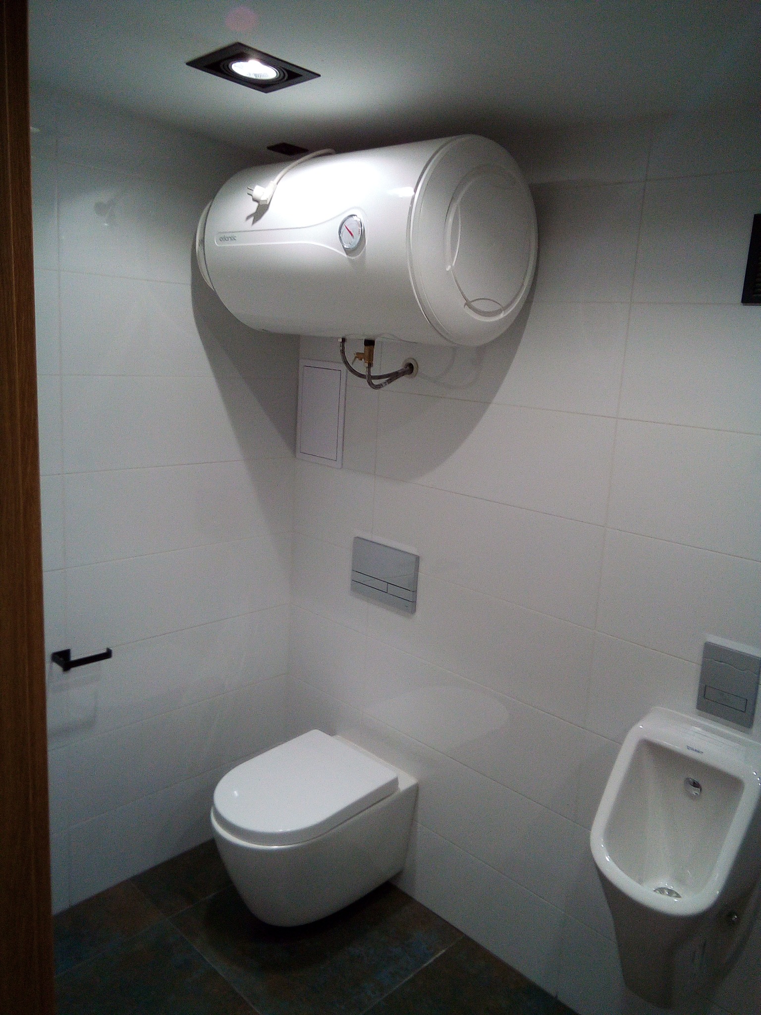 Biała łazienka z podwieszanym bojlerem Atlantic nad toaletą i pisuar obok, wykończona białymi płytkami i ciemną podłogą, oświetlona punktowym światłem.