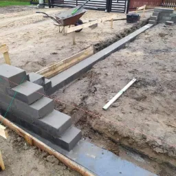 Wykop z betonową wylewką fundamentową, częściowo obudowany drewnianym szalunkiem, z fragmentem murowanej, schodkowej konstrukcji z bloczków betonowych, poziomica i rury PCV na ziemi, taczka w tle.