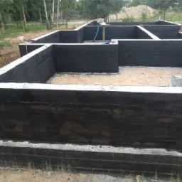 Fundamenty domu murowanego z hydroizolacją bitumiczną, widoczne zbrojenia i teren budowy z drzewami w tle.