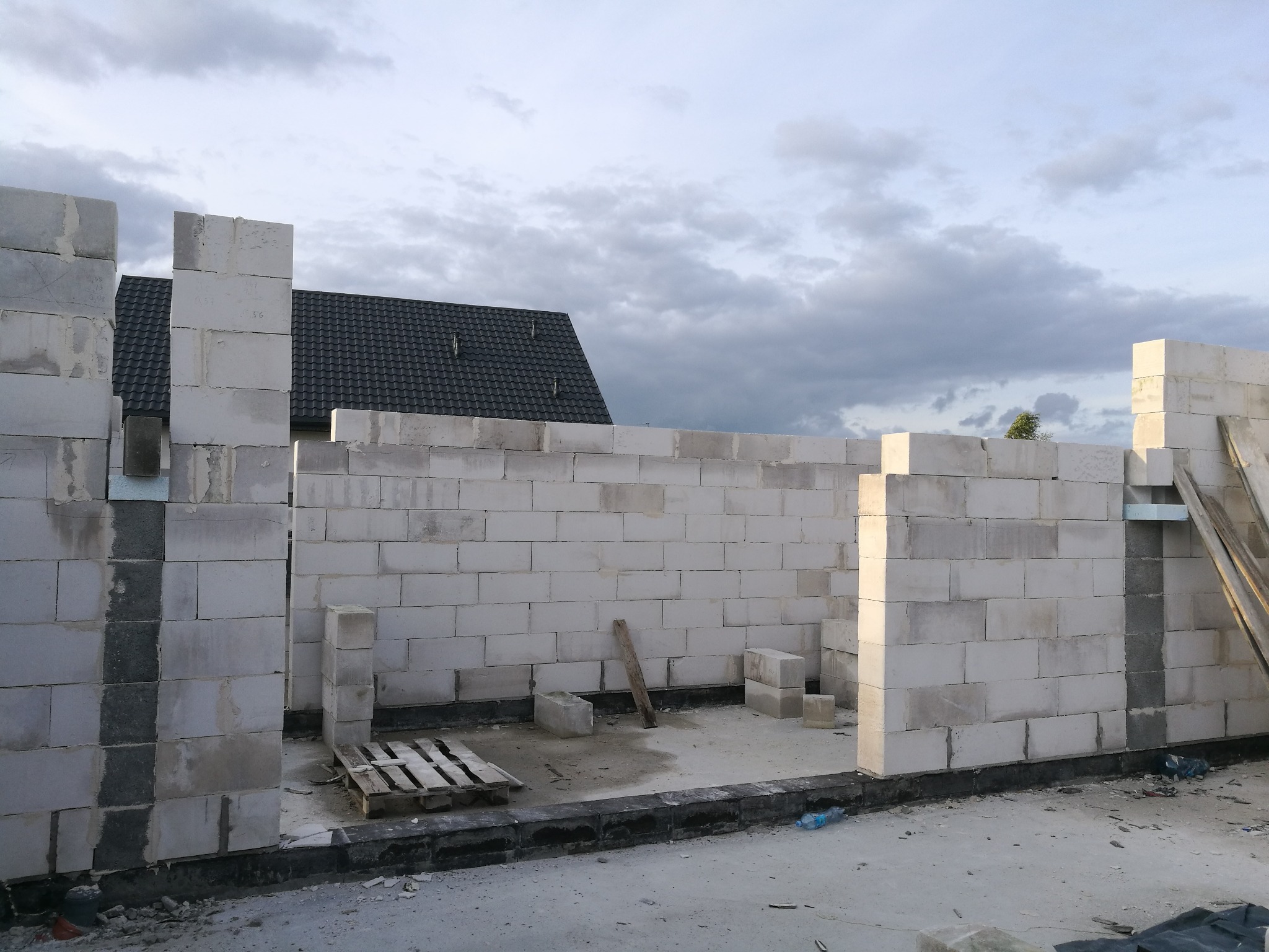 Ściany budynku w trakcie wznoszenia z białych bloczków, widoczne fragmenty dachu i niebo z chmurami w tle.