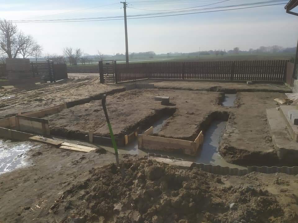 Przygotowanie fundamentów pod budowę domu: wykopane rowy zalane betonem, drewniane szalunki w ziemi, łopata wbita w ziemię, widok na ogrodzenie i krajobraz w tle.