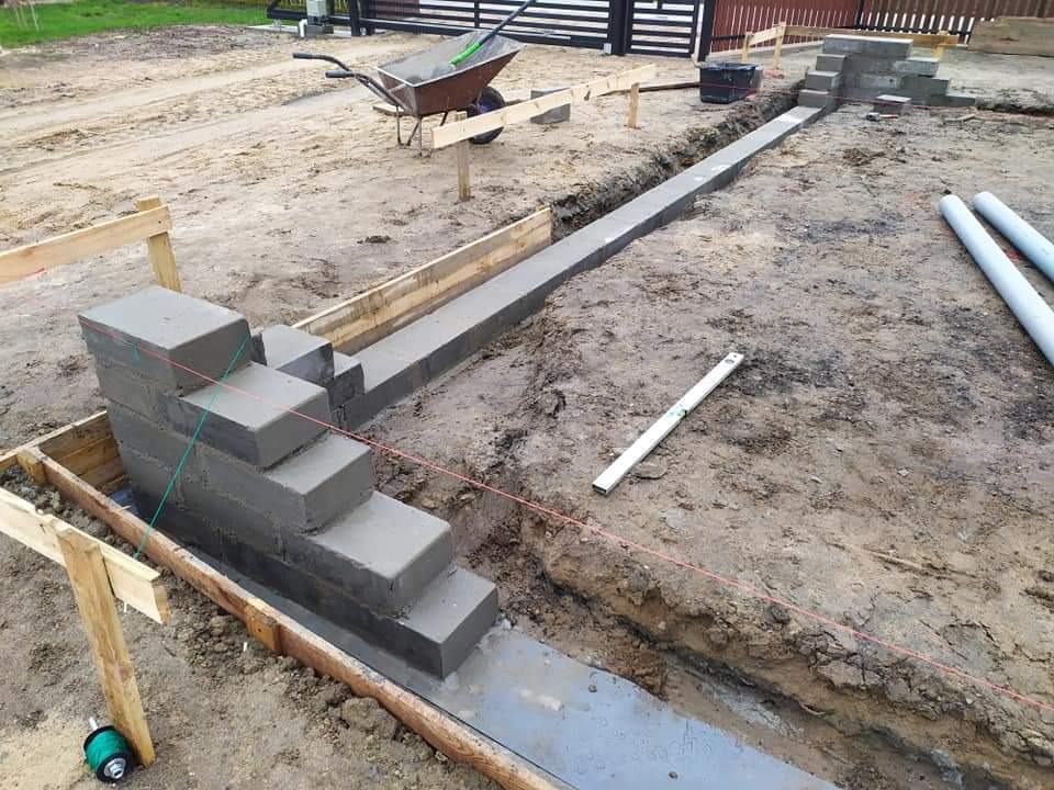 Wykop z betonową wylewką fundamentową, częściowo obudowany drewnianym szalunkiem, z fragmentem murowanej, schodkowej konstrukcji z bloczków betonowych, poziomica i rury PCV na ziemi, taczka w tle.