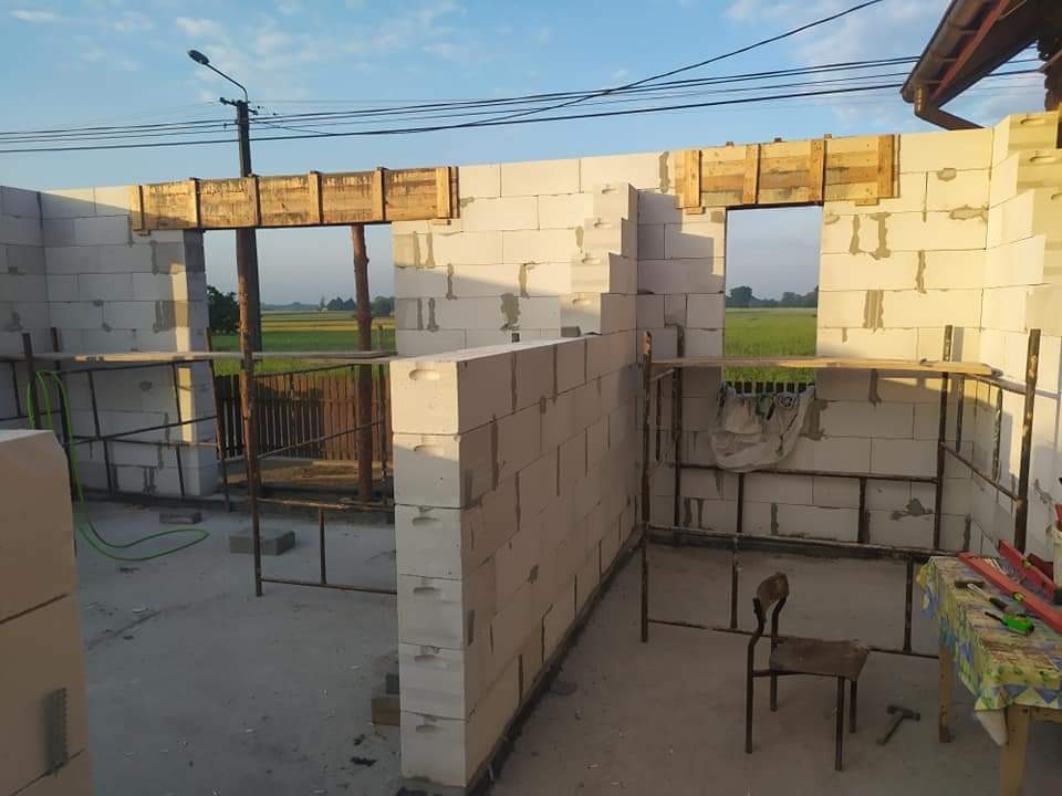 Wznoszenie ścian z betonu komórkowego Ytong w trakcie budowy domu jednorodzinnego, widoczne drewniane nadproża okienne i drzwiowe, rusztowania i narzędzia na budowie, pole w tle.