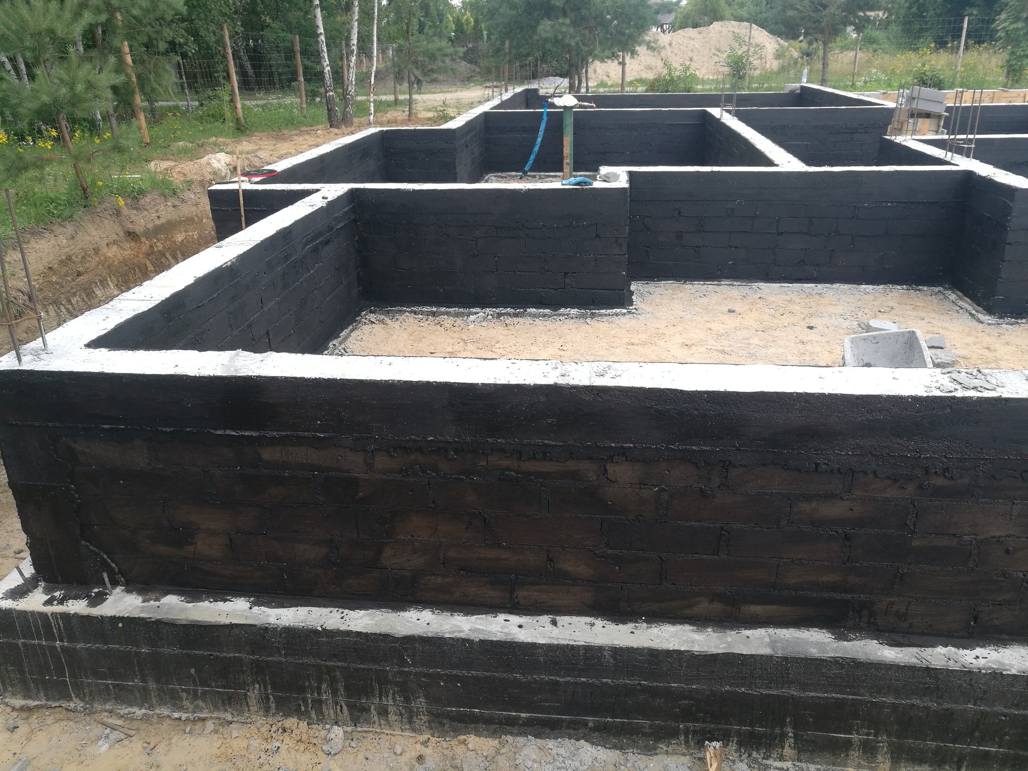 Fundamenty domu murowanego z hydroizolacją bitumiczną, widoczne zbrojenia i teren budowy z drzewami w tle.