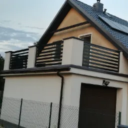 Dwukondygnacyjny dom z balkonem zabezpieczonym czarną, poziomą balustradą metalową; widoczna brama garażowa i siatka ogradzająca posesję.