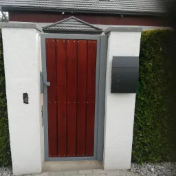 Drewniana furtka w metalowej ramie, wkomponowana w białe, otynkowane słupki, z czarną skrzynką na listy, przy posesji w Szczecinie, otoczona zielonym żywopłotem.