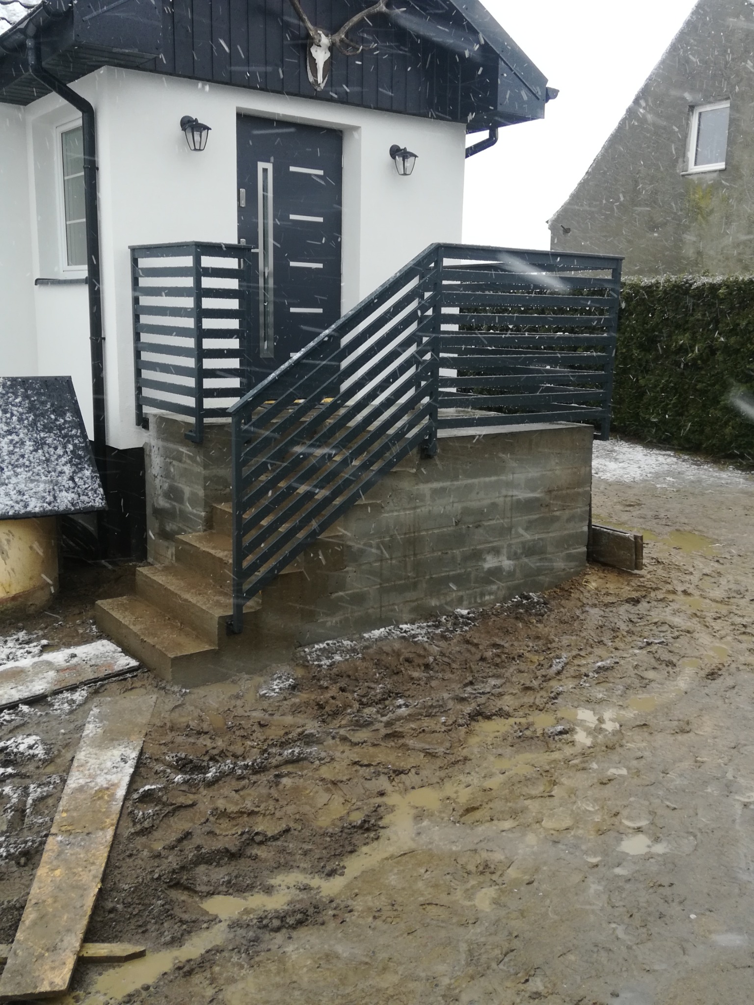 Metalowe barierki schodowe z poziomymi szczebelkami prowadzące do drzwi wejściowych domu w zimowej scenerii, z błotnistym podłożem i padającym śniegiem.