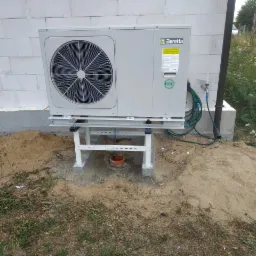 Zamontowana pompa ciepła Beretta na metalowej konstrukcji przy ścianie z bloczków, widoczny fragment instalacji hydraulicznej i zielony wąż.