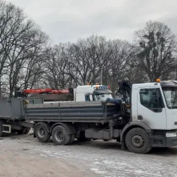 Ciężar&oacute;wka z przyczepą i zamontowanym dźwigiem hydraulicznym, zaparkowana na placu z widocznymi drzewami w tle, w pochmurny dzień.