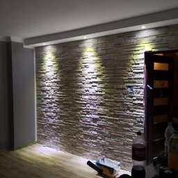 Total Kwadrat - Ściana wykończona dekoracyjną okładziną kamienną z podświetleniem punktowym, widok na fragment podłogi i akcesoria malarskie na stole. Efektowna aranżacja.