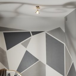 Total Kwadrat - Ściana w pokoju z geometrycznym wzorem w odcieniach szarości i bieli, oświetlona dwoma punktowymi lampami sufitowymi, fragment łóżka z ubraniami.