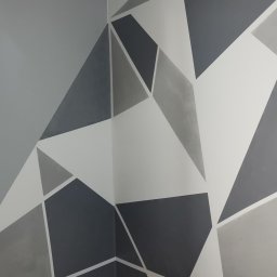 Total Kwadrat - Geometryczny wzór na ścianie w odcieniach szarości i bieli, widok na róg pokoju, drewniana podłoga, narzędzia malarskie w rogu. Dekoracyjne malowanie ścian.
