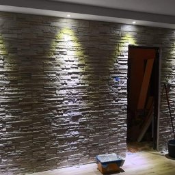 Total Kwadrat - Ściana wykończona dekoracyjną okładziną imitującą kamień, oświetlona punktowymi lampami sufitowymi. Widoczne narzędzia i akcesoria malarskie.