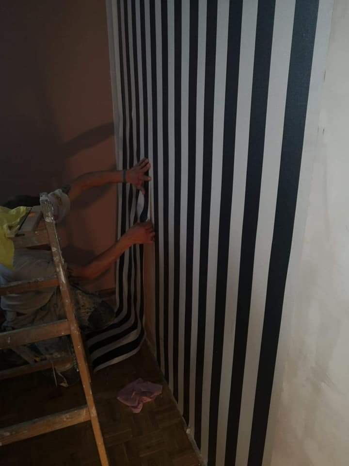 Pracownik na drewnianej drabinie nakleja tapetę w czarno-białe pionowe pasy na ścianie w rogu pokoju, widoczny fragment podłogi z leżącą szmatką.