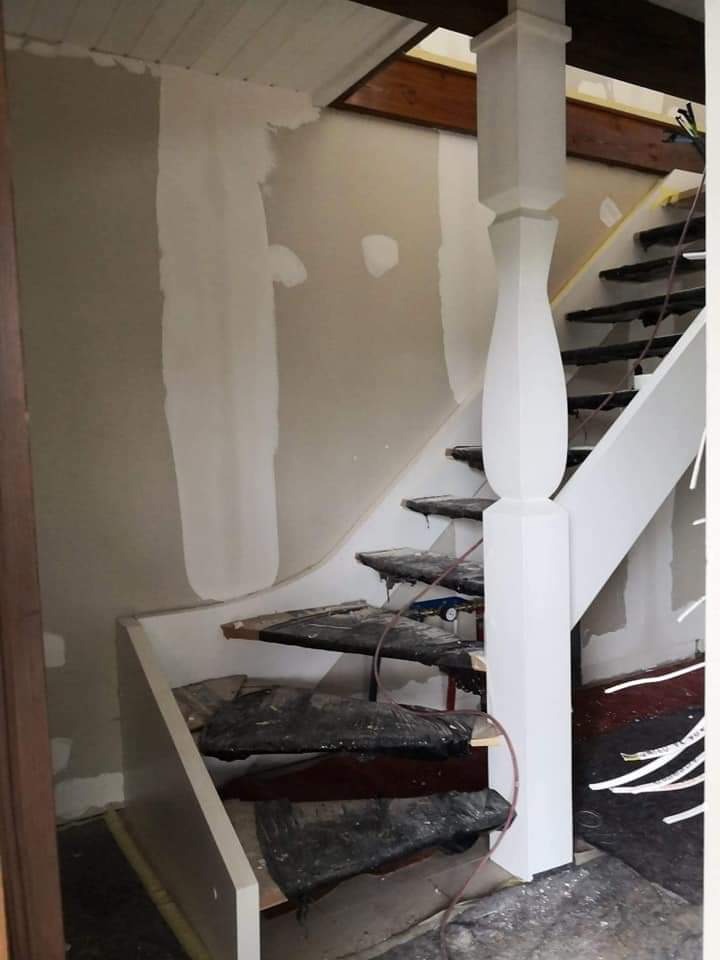 Klatka schodowa w trakcie prac wykończeniowych: ściany po wstępnym wyrównaniu, schody zabezpieczone folią, widoczny drewniany strop.