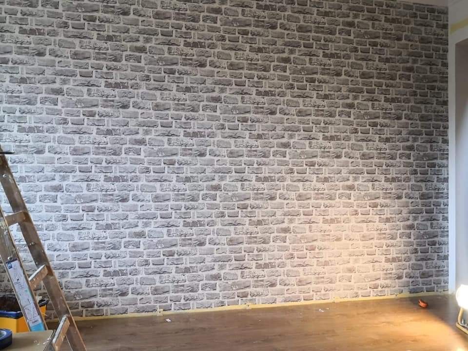 Ściana wykończona tapetą imitującą szarą cegłę, widoczna drabina malarska, podłoga zabezpieczona taśmą malarską.