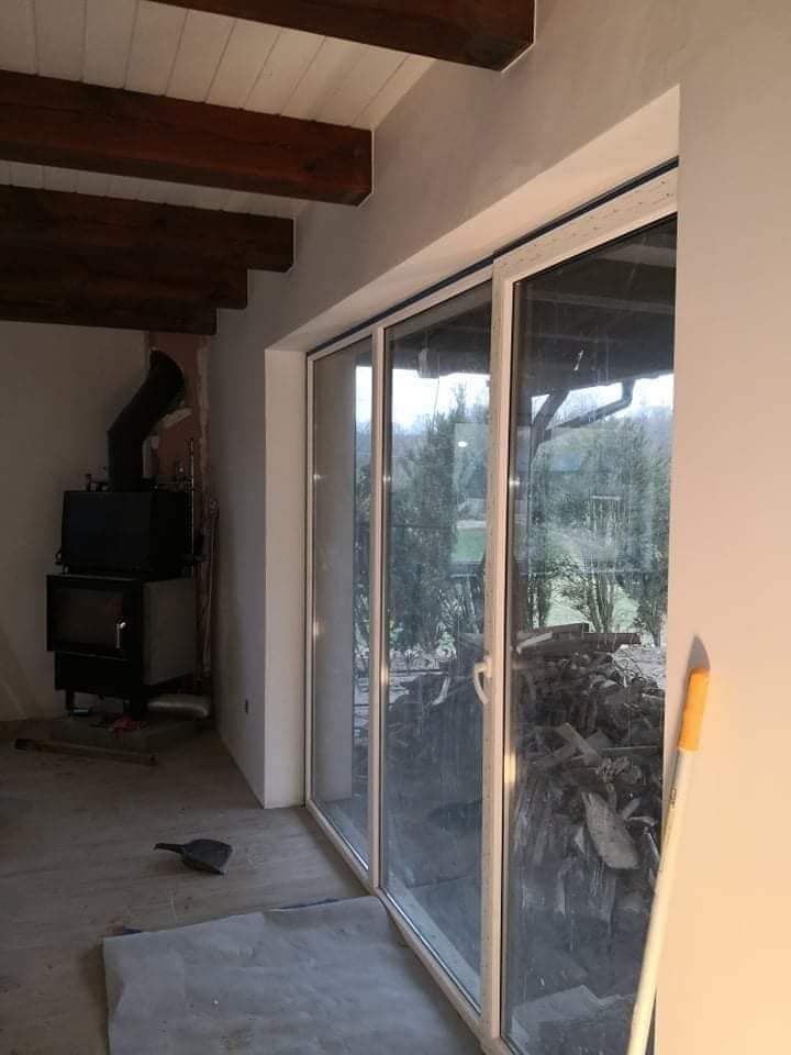 Wnętrze w trakcie remontu z widocznym piecem, drewnianymi belkami stropowymi i dużymi przesuwnymi drzwiami balkonowymi z widokiem na zewnątrz, podłoga zabezpieczona folią malarską.
