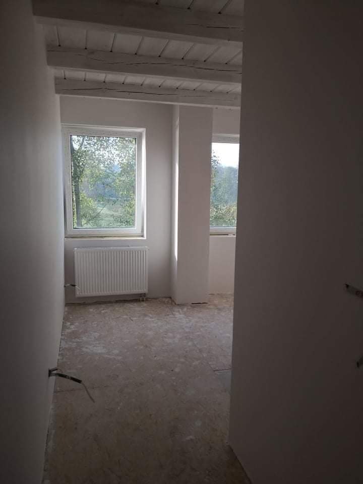 Wnętrze pokoju w trakcie prac wykończeniowych: ściany po nałożeniu gładzi, widoczne dwa okna z widokiem na zieleń, biały grzejnik, drewniane belki stropowe, podłoga przygotowana do dalszych prac.