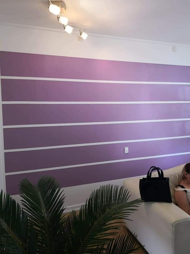 Fioletowa ściana z poziomymi białymi pasami, oświetlona lampami sufitowymi, z częściowo widoczną sofą i rośliną doniczkową na pierwszym planie.