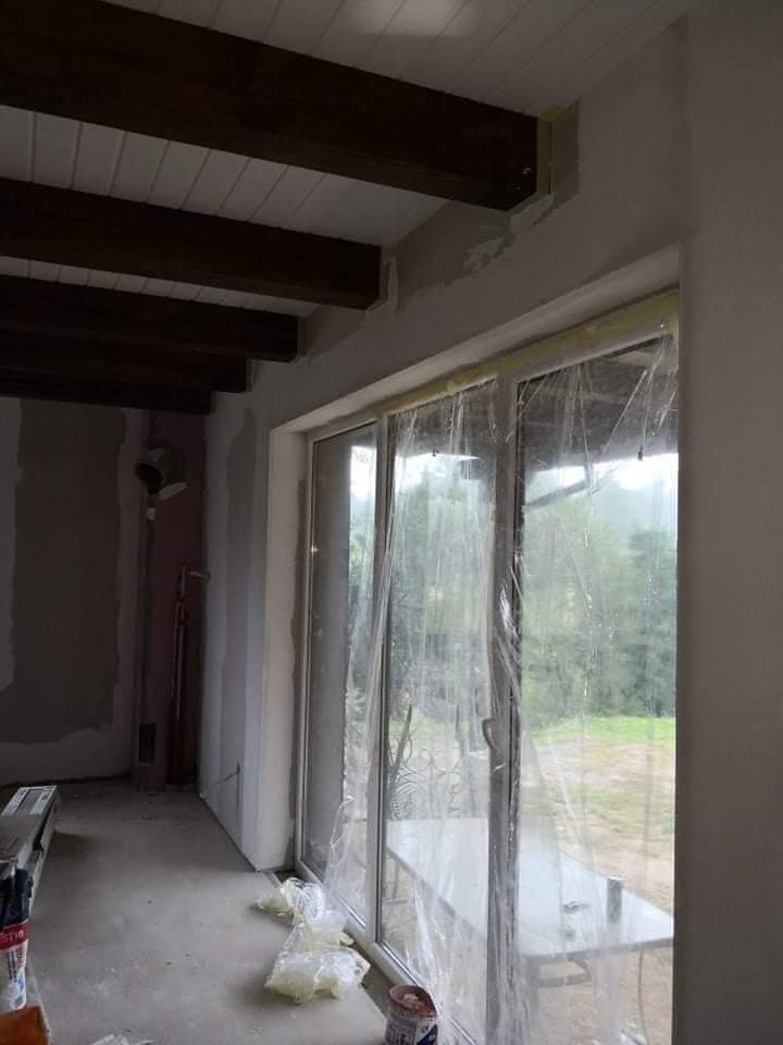 Wnętrze w trakcie prac wykończeniowych: surowy beton na podłodze, ściany przygotowane pod malowanie, okno zabezpieczone folią, widoczne drewniane belki stropowe.
