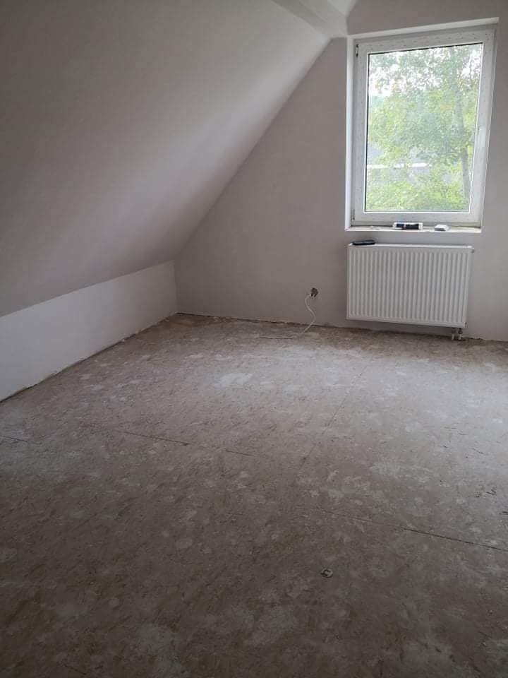 Poddasze po wstępnym przygotowaniu ścian, widoczne ślady po pracach na podłodze, okno z widokiem na drzewa, grzejnik pod oknem.