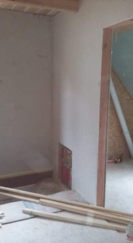 Wnętrze w trakcie prac wykończeniowych: surowe ściany, drewniane elementy konstrukcyjne widoczne przy podłodze i suficie, fragment otworu drzwiowego z drewnianą ościeżnicą.