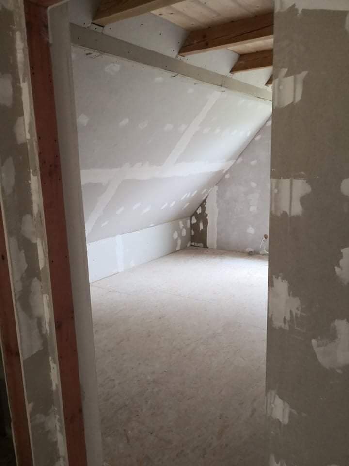 Poddasze w trakcie wykańczania: widoczne płyty karton-gips na skosach, drewniane belki stropowe i surowe ościeżnice drzwiowe, ślady szpachlowania na ścianach.