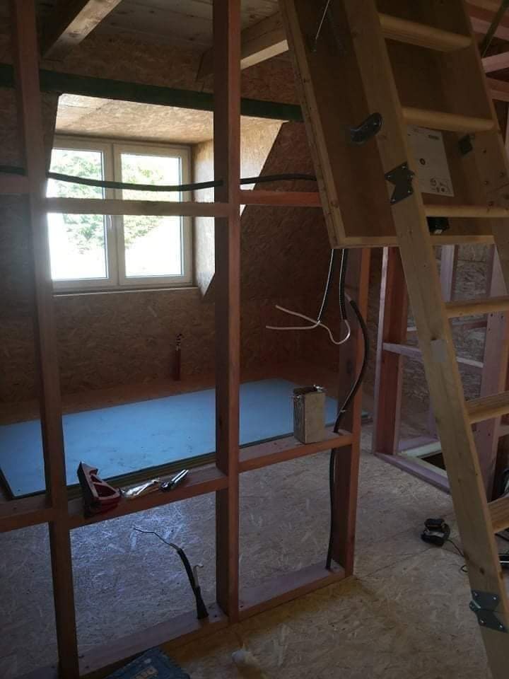 Wnętrze poddasza w trakcie adaptacji, widoczne drewniane rusztowanie, okno dachowe, drabina i narzędzia budowlane. Ściany wykończone płytą OSB.