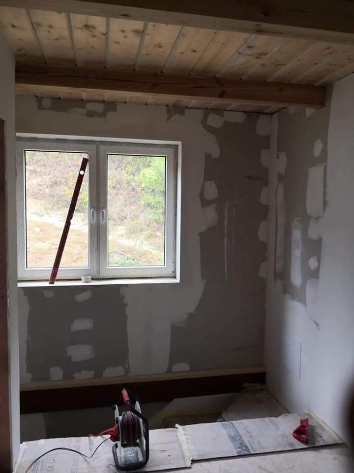 Wnętrze pokoju w trakcie prac wykończeniowych: częściowo zagruntowane ściany z widocznymi łatami, drewniany sufit, poziomica oparta o okno, przedłużacz na podłodze.