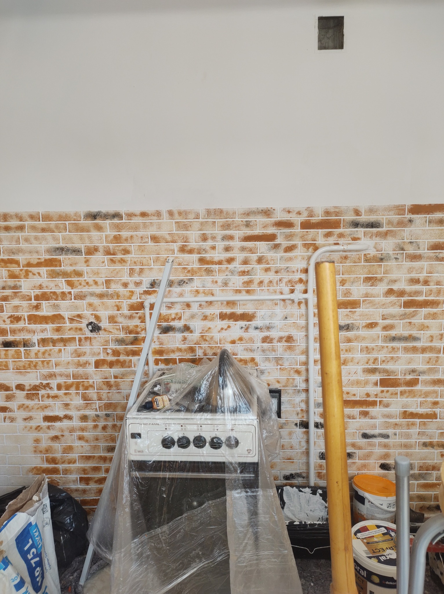 Kuchenka zabezpieczona folią w trakcie prac. Tło stanowi ściana z cegieł pomalowana na biało, widoczne narzędzia malarskie i puszki z farbą.
