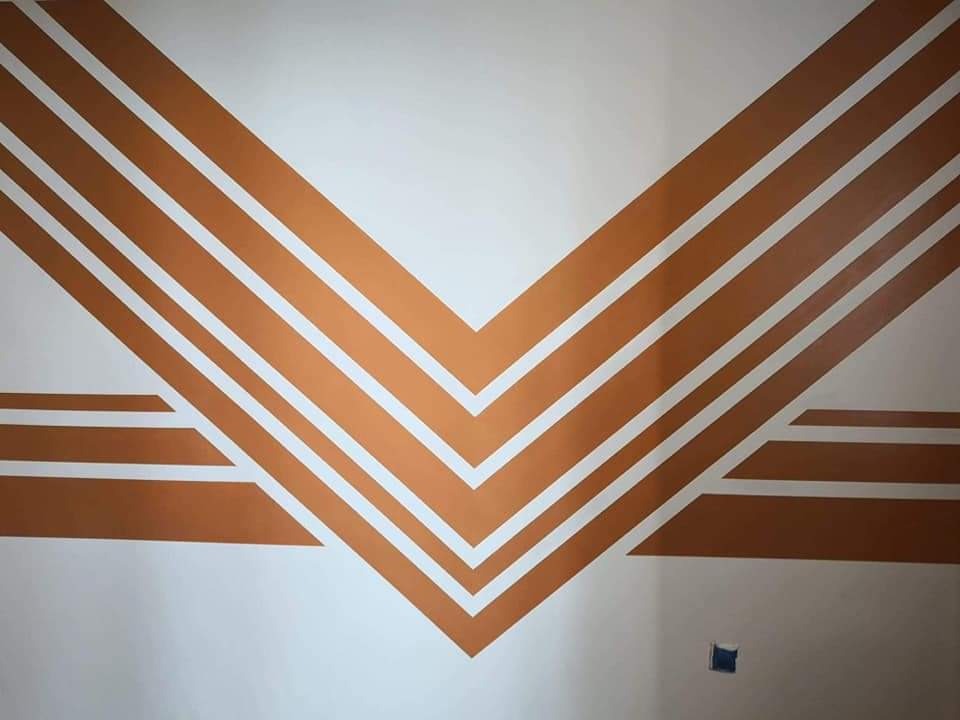 Biała ściana z dekoracyjnym, geometrycznym wzorem w kolorze miedzianym. Wzór tworzą równoległe linie układające się w kształt litery V i poziome pasy.
