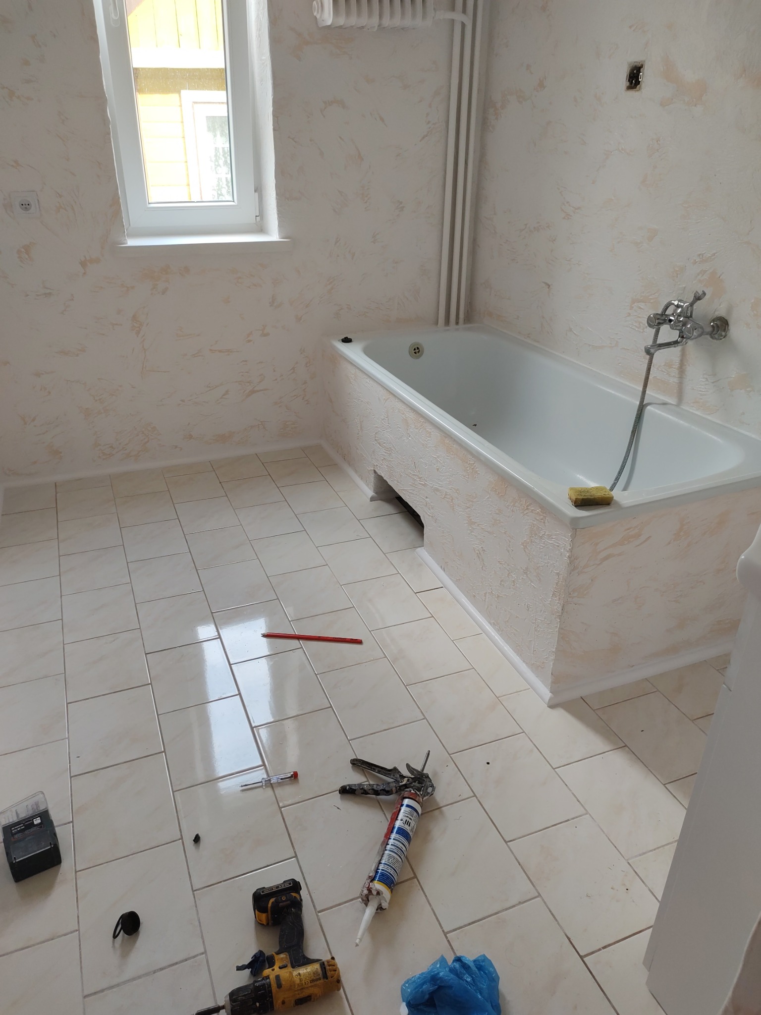 Wykończona łazienka z wanną obudowaną dekoracyjną strukturą, narzędzia na podłodze z płytek. Ściany z efektem dekoracyjnym, okno z widokiem na żółty budynek.