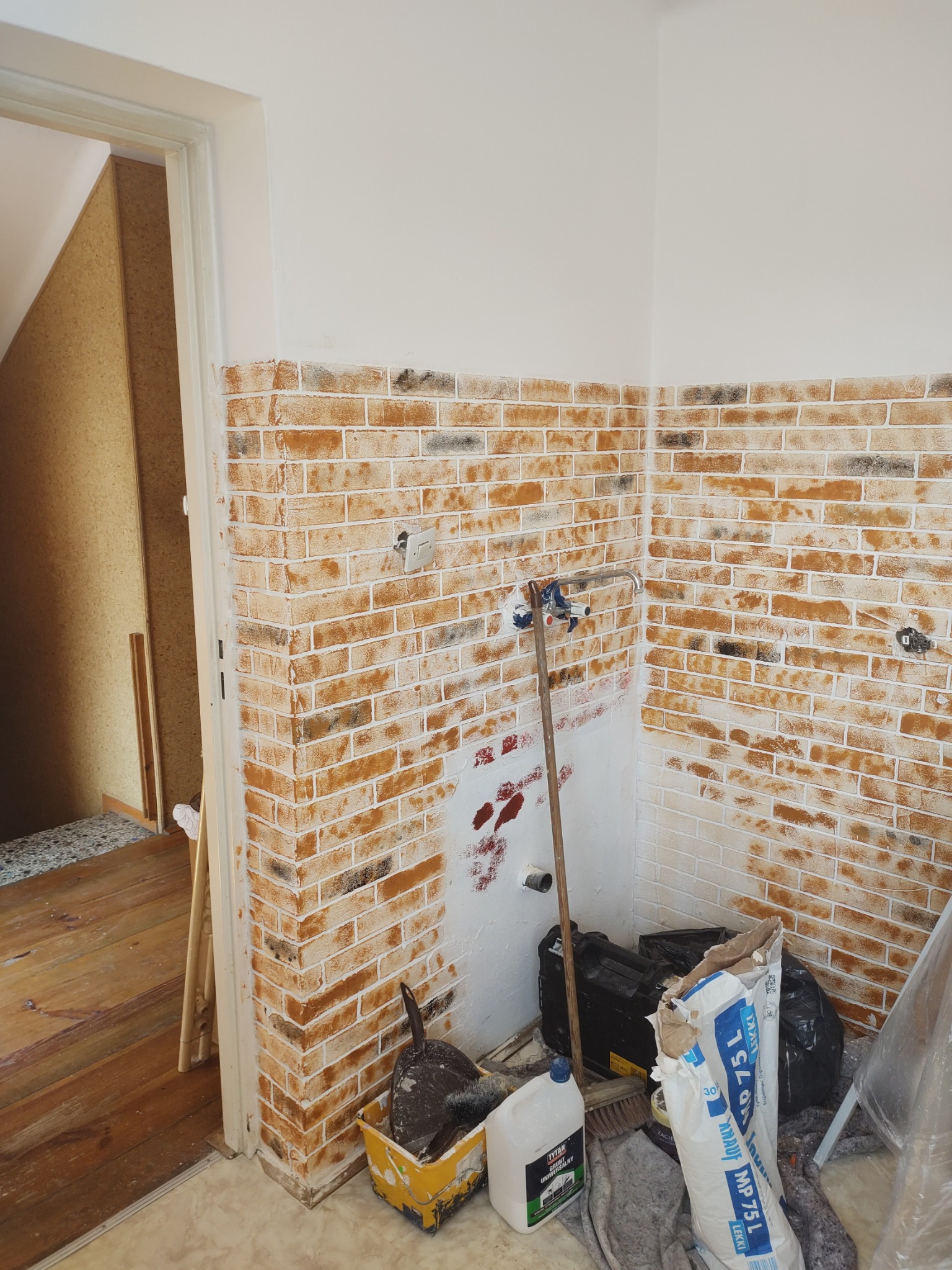 Ściana z imitacją cegły w trakcie wykańczania, z widocznymi narzędziami i materiałami budowlanymi. Częściowo pomalowana na biało, z instalacją hydrauliczną na ścianie.