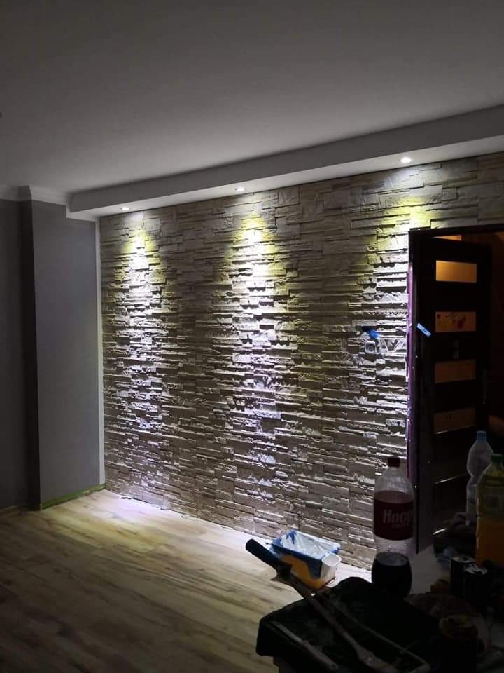 Ściana wykończona dekoracyjną okładziną kamienną z podświetleniem punktowym, widok na fragment podłogi i akcesoria malarskie na stole. Efektowna aranżacja.