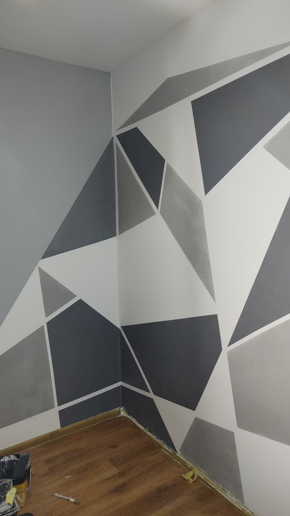 Geometryczny wzór na ścianie w odcieniach szarości i bieli, widok na róg pokoju, drewniana podłoga, narzędzia malarskie w rogu. Dekoracyjne malowanie ścian.