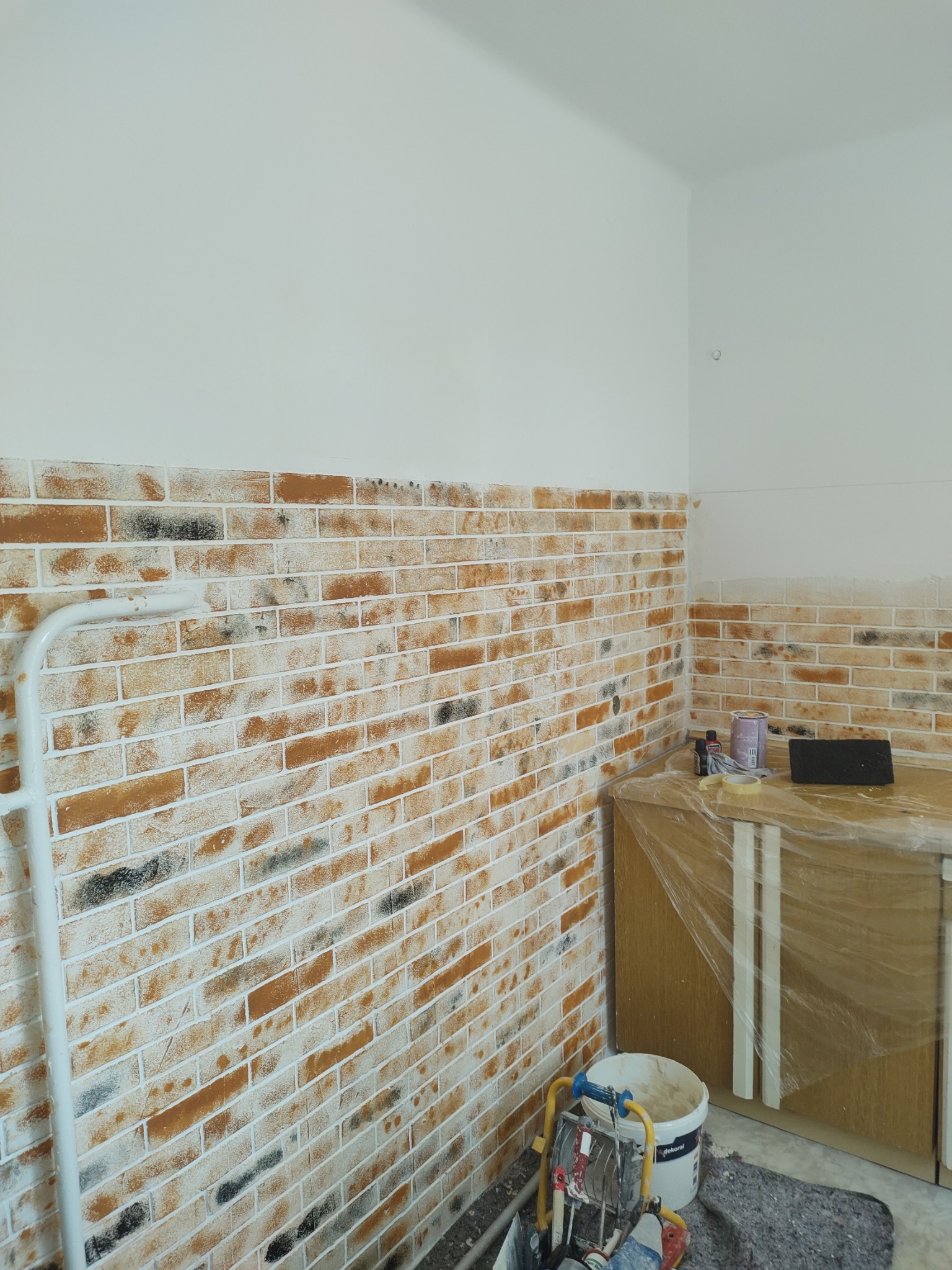 Ściana z efektem cegły, częściowo pomalowana na biało, z widocznymi narzędziami malarskimi i zabezpieczonymi meblami, sugerująca etap prac wykończeniowych.