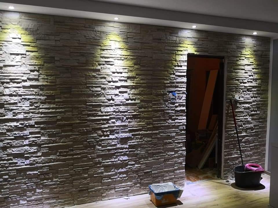 Ściana wykończona dekoracyjną okładziną imitującą kamień, oświetlona punktowymi lampami sufitowymi. Widoczne narzędzia i akcesoria malarskie.