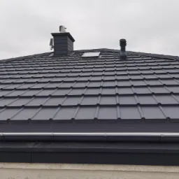 Szary dach z blachy trapezowej z kominem, odpowietrznikiem i oknem dachowym, widok z dołu, pochmurne niebo w tle.