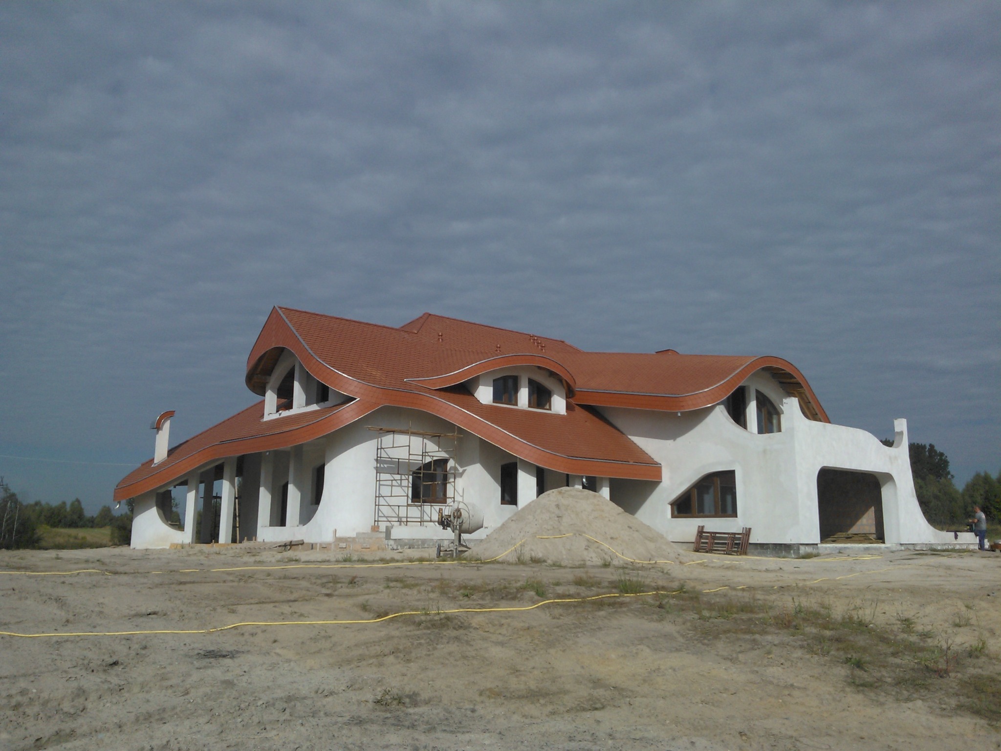 Niedokończony dom z falistym dachem z czerwonej dachówki, widoczne rusztowania, mieszalnik do betonu i sterta piasku na placu budowy.