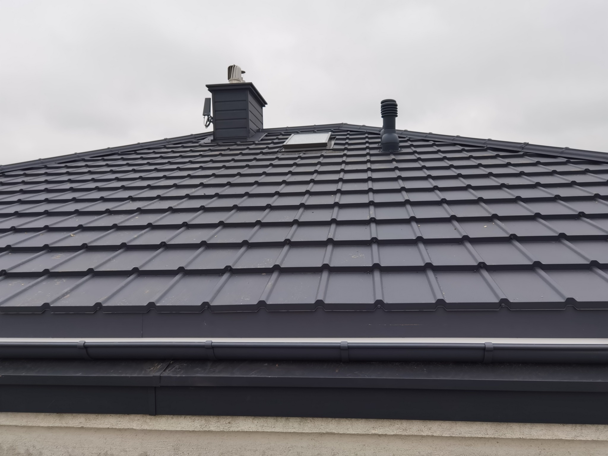Szary dach z blachy trapezowej z kominem, odpowietrznikiem i oknem dachowym, widok z dołu, pochmurne niebo w tle.