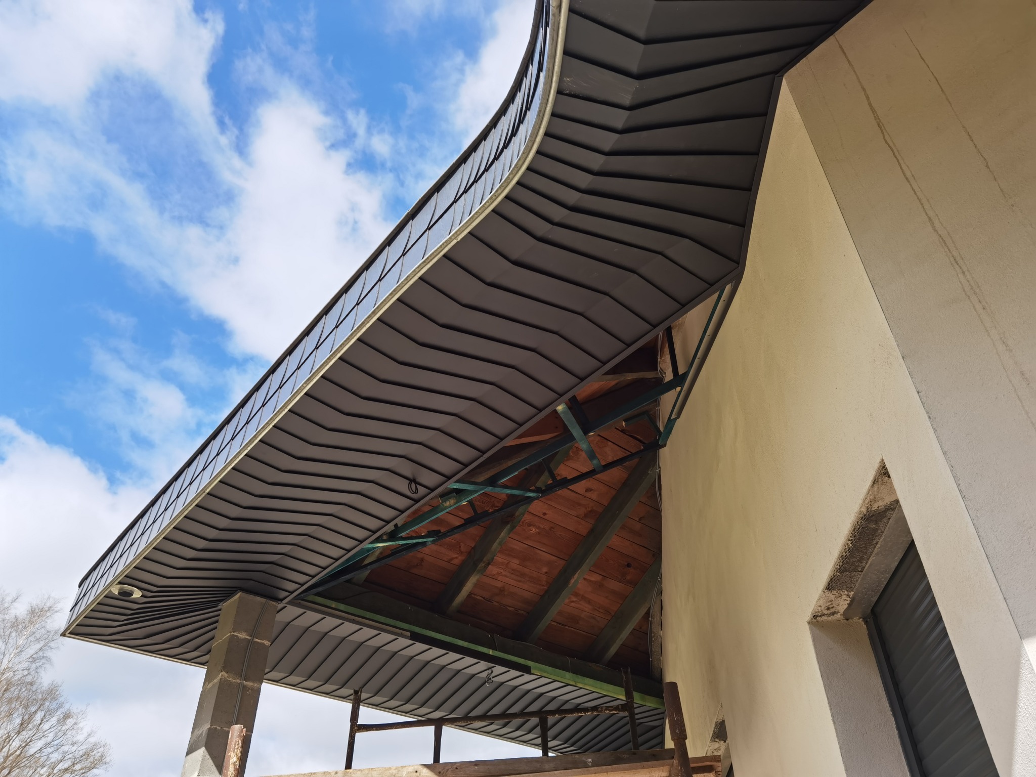 Nowoczesna konstrukcja dachu z szarej blachy trapezowej, widok z dołu na drewnianą więźbę i fragment elewacji budynku w trakcie wykończenia, na tle błękitnego nieba z chmurami.