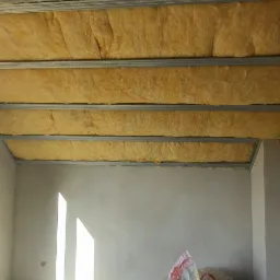 Poddasze w trakcie ocieplania, widoczna wełna mineralna między metalowymi profilami konstrukcyjnymi, na podłodze resztki materiałów i worek ze śmieciami, promienie słoneczne wpadają przez okno.