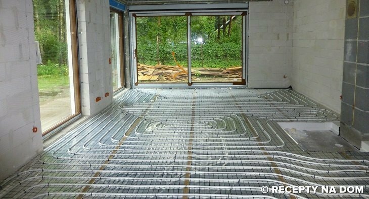 Instalacja ogrzewania podłogowego w surowym stanie, widoczne rury ułożone na siatce, ściany z betonu komórkowego, duże okno z widokiem na zewnątrz.