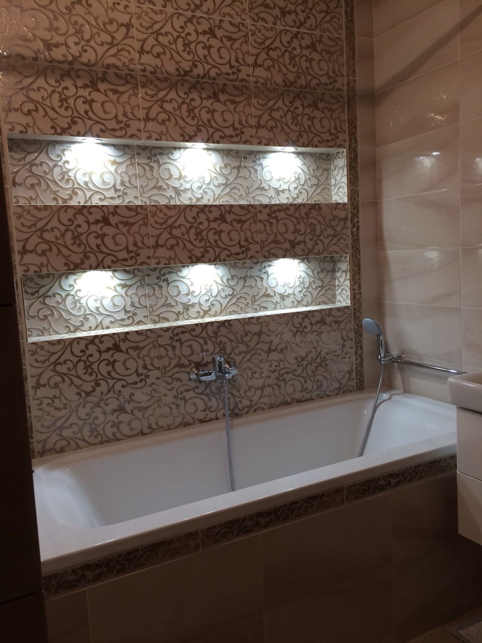 Wykończona łazienka z wanną, baterią, i dekoracyjnymi, podświetlanymi wnękami w ścianie z płytek o wzorze ornamentowym.