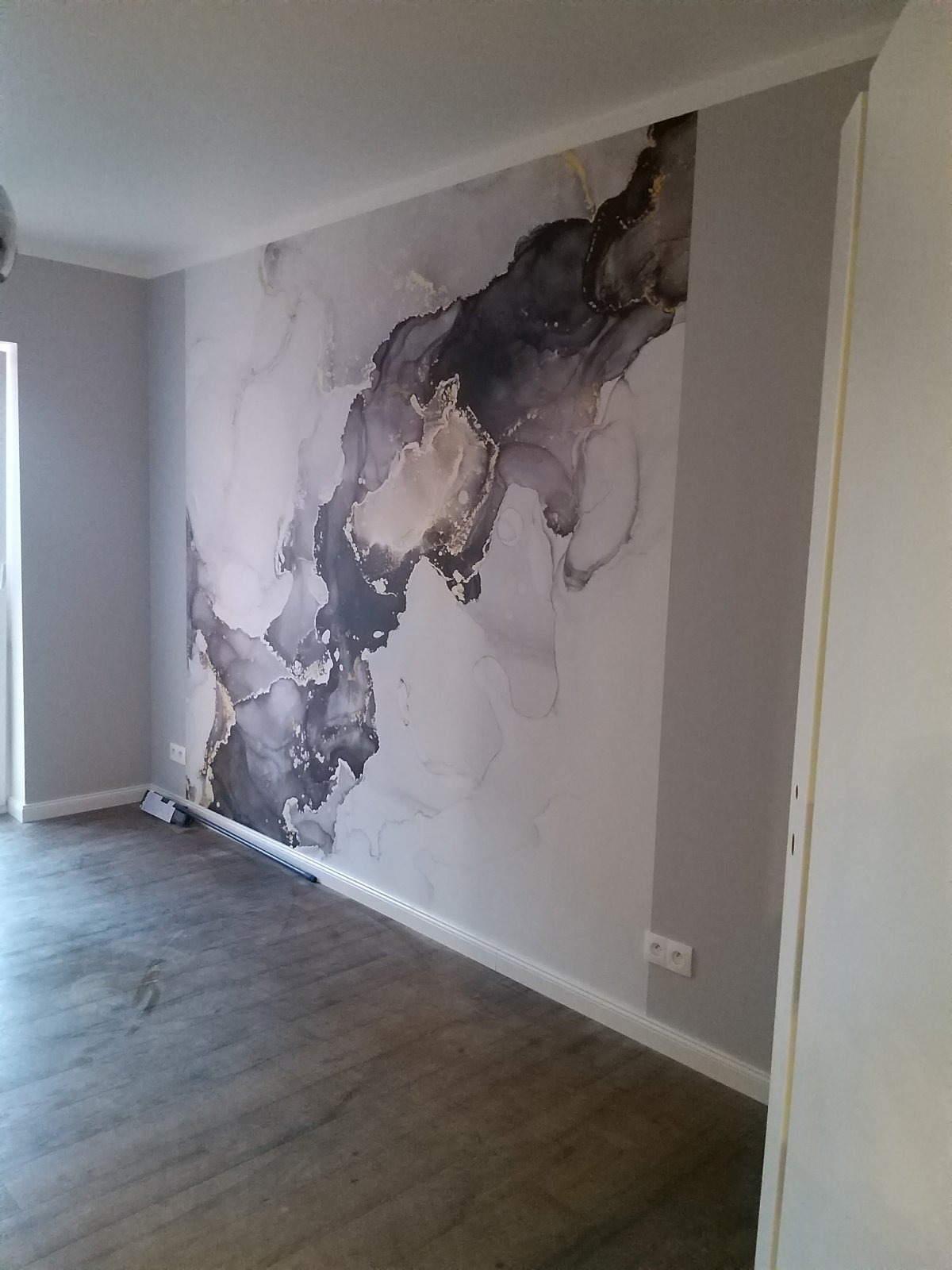 Ściana pokoju z abstrakcyjną tapetą w odcieniach szarości, bieli i złota, pasująca do szarych ścian i podłogi, z listwą przypodłogową i gniazdkami elektrycznymi.