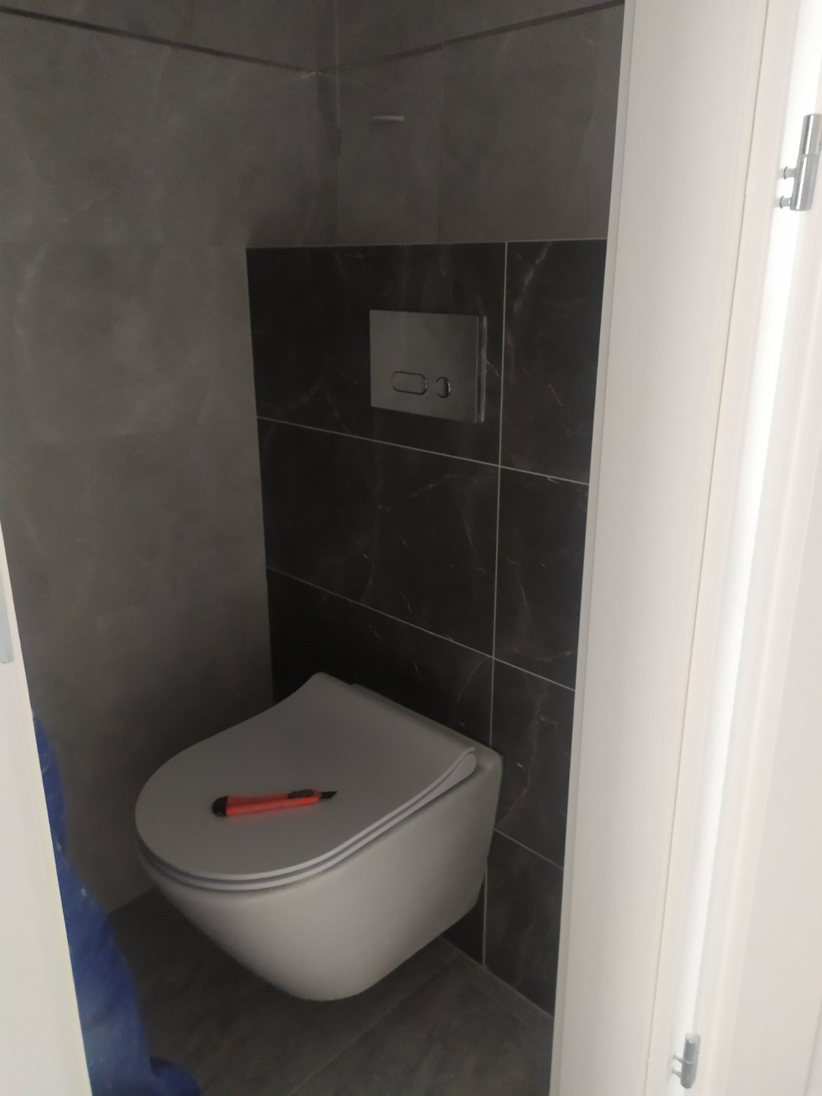 Zainstalowana podwieszana toaleta z przyciskiem spłukującym wkomponowanym w ciemne płytki imitujące marmur, na desce toalety leży czerwony nożyk introligatorski.