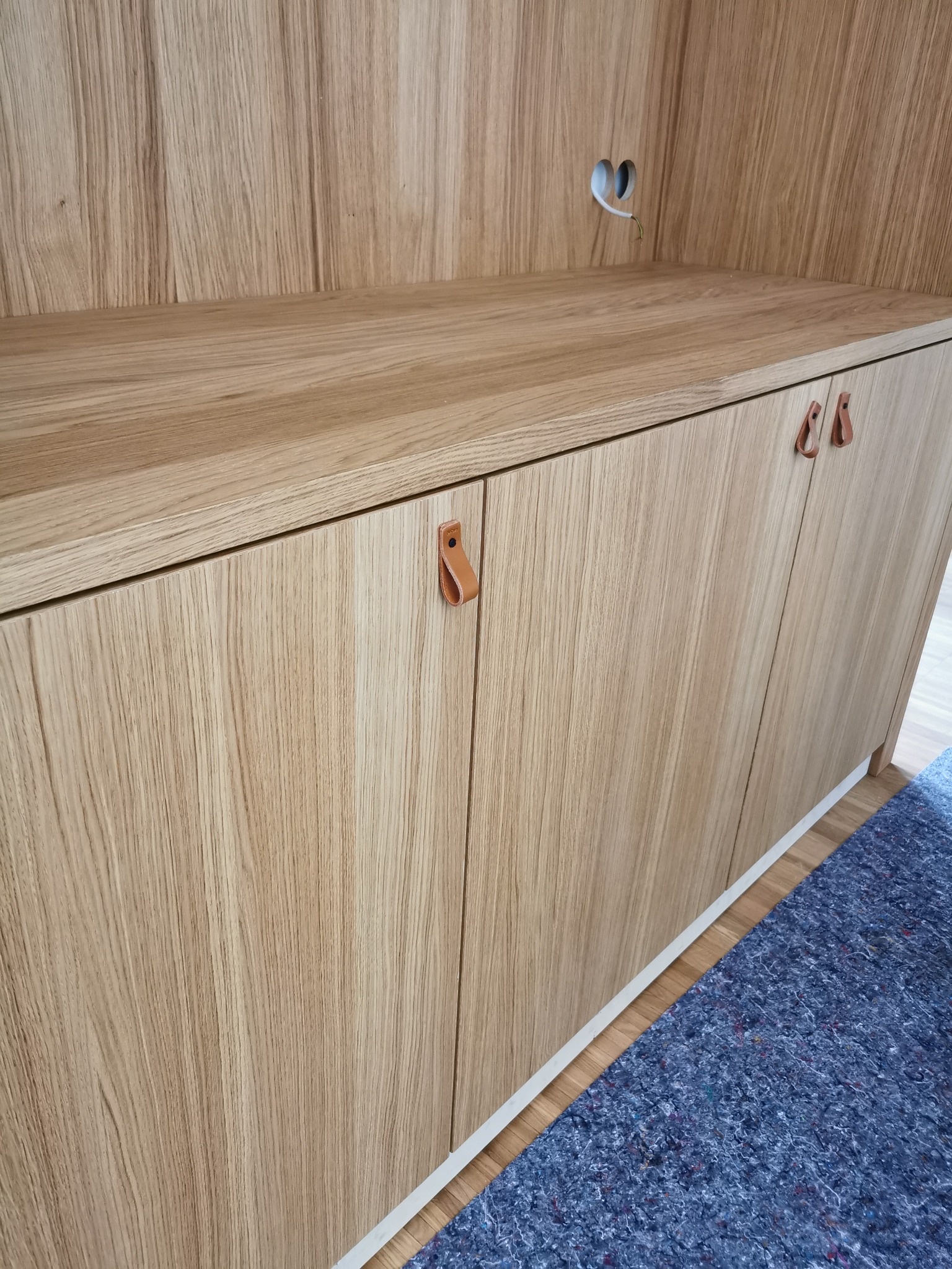 Drewniana zabudowa szafkowa z jasnego drewna z widocznymi skórzanymi uchwytami i gniazdkiem elektrycznym w górnej części.