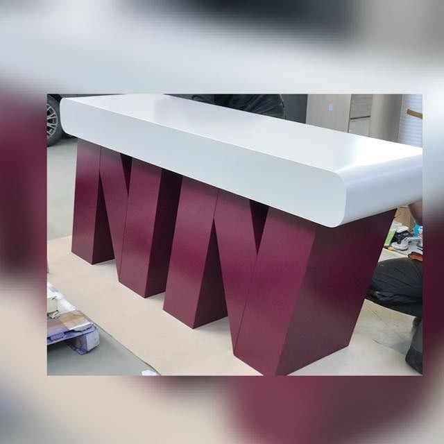 Biały blat lady recepcyjnej wsparty na bordowych, geometrycznych nogach w kształcie litery N, widoczne częściowo elementy otoczenia.