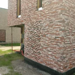 Fragment ściany zewnętrznej budynku z czerwonej cegły z otworami wentylacyjnymi, widoczne okno bez ramy i podpora tymczasowa.